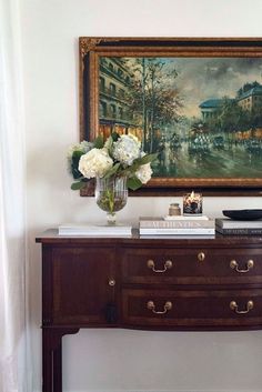 a painting hangs on the wall above a dresser with books and flowers in front of it