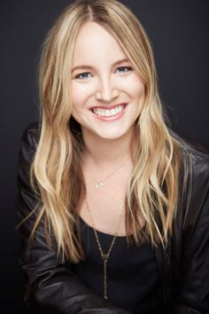 a woman with long blonde hair wearing a black jacket and smiling at the camera in front of a dark background