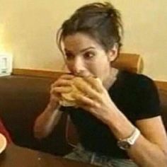 a woman sitting at a table eating a doughnut in front of her face with both hands