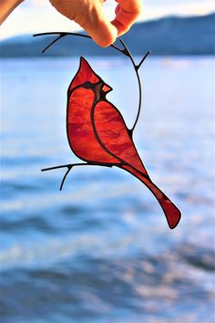 a hand holding a stained glass red bird on a branch by the water's edge