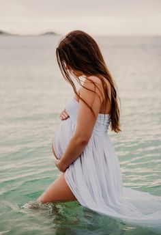 a pregnant woman standing in the ocean with her belly wrapped around her waist and wearing a white dress