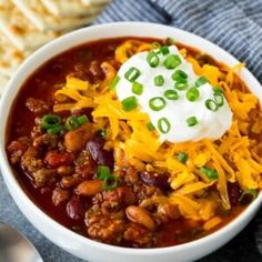 a white bowl filled with chili and cheese next to crackers on a blue towel