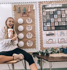 a woman sitting in front of a bulletin board holding a coffee cup and smiling at the camera