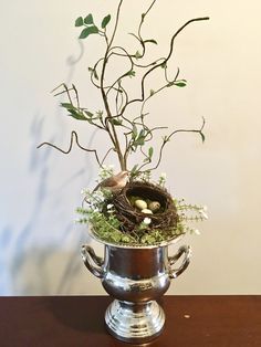 a bird's nest in a silver vase filled with greenery on a table