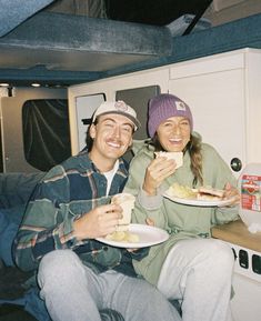 two people eating food in the back of a vehicle on a road trip, one man is holding a plate and smiling at the camera