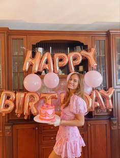 a woman standing in front of a birthday cake with balloons on the wall behind her