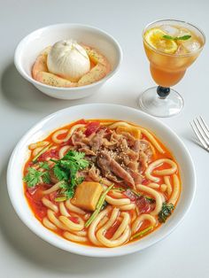 a bowl of noodles, meat and vegetables next to a glass of orange juice on a white table