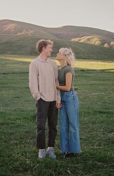 a man and woman standing in the grass with mountains in the background