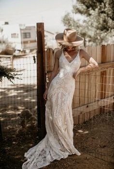 a woman in a white dress and cowboy hat leaning against a fence with her back to the camera