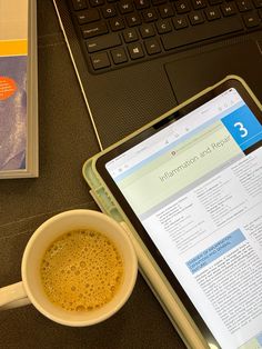 a cup of coffee sitting next to a laptop on a desk with an open book