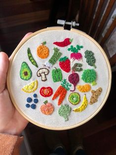 a person holding up a small embroidery hoop with fruits and vegetables on it in front of a window