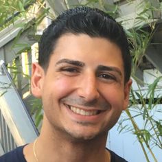 a man with a moustache smiles at the camera while standing in front of some plants