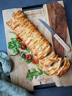 a wooden cutting board topped with meat covered in cheese and vegetables next to a knife