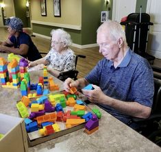 an older man and woman playing with blocks at a table in a nursing home while others look on