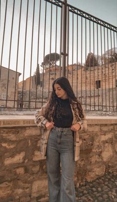 a woman standing in front of a stone wall with her hands on her hips and looking off into the distance