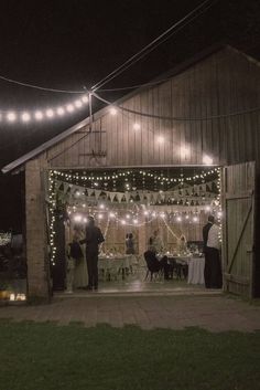 people standing in front of a barn at night with lights strung from the roof and around them