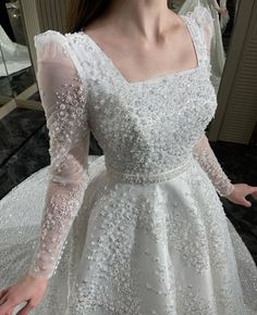 a woman wearing a white dress with long sleeves and beading on the skirt is standing in front of a mirror