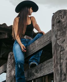 a woman sitting on top of a wooden fence wearing jeans and a black cowboy hat