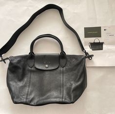 "Longchamp 100% AUTHENTIC Le Pliage Cuir Leather Shoulder Bag with Logo Strap, Black Leather with Gunmetal Hardware, Original $630 Women or Men can use it too  Rich leather and impeccable topstitching give a refined aesthetic to Longchamp's iconic Le Pliage shoulder bag, branded with an equestrian logo. Slim top handles and a removable wide logo shoulder strap let you style the chic look you prefer. Top-zip closure with snap-flap tab Top handles; removable, adjustable shoulder strap Interior zip Black Leather Bag Aesthetic, Black Leather Travel Bag, Longchamp Leather Bag, Longchamp Cuir, Leather Longchamp, Equestrian Logo, Designer Handbags Louis Vuitton, Longchamp Leather, Black Duffel Bag