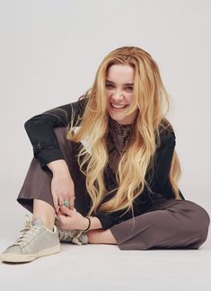 a woman with long blonde hair sitting on the ground smiling and holding her hand out