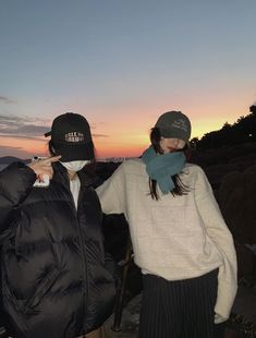 two people standing next to each other in front of the sun at dusk with one person wearing a hat and scarf