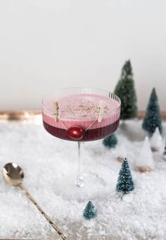 a drink in a martini glass with a cherry on the rim and small trees behind it