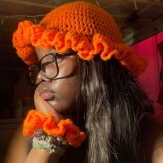 a woman wearing an orange crocheted hat and glasses with her hand on her chin