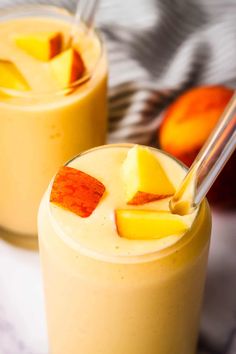 two glasses filled with orange smoothie on top of a white table next to sliced peaches