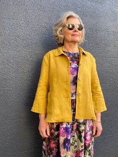 an older woman standing in front of a gray wall wearing yellow jacket and floral pants