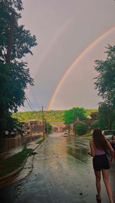 a woman walking down a rain soaked street with a rainbow in the sky behind her