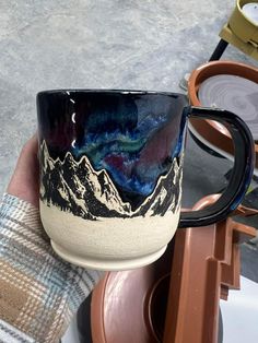 a hand holding a black and white coffee cup with mountains on the side, in front of other items