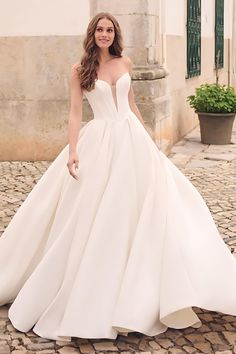 a woman in a white wedding dress standing on a cobblestone street with her hands behind her back