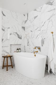 a white bath tub sitting next to a wooden stool in a bathroom with marble walls and floors