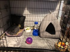 a cage filled with food and other items including a cat bed, water bottle and bowl