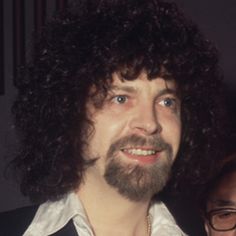 a man with long curly hair and glasses smiling at the camera while standing next to another man