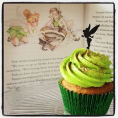 a cupcake with green frosting and a tinkerbell on top sits in front of an open book