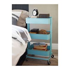 a blue cart with books and an alarm clock on top is in front of a bed