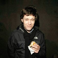 a man in a black jacket is eating something out of his hand and looking at the camera