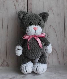 a gray and white crocheted cat sitting on top of a wooden table with a pink ribbon around its neck