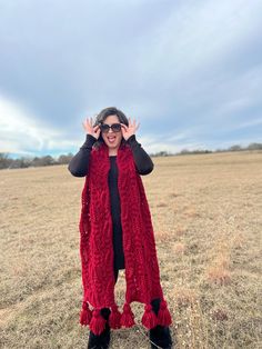 a woman standing in a field wearing a red knitted scarf and holding her hands up