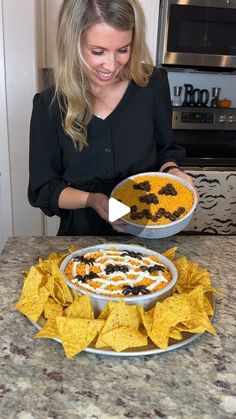 a woman standing in front of a bowl of food with chips on the table next to it