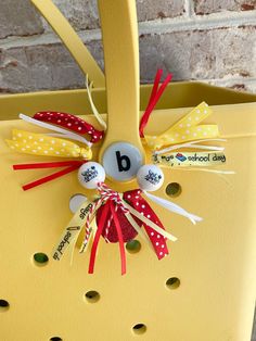 a close up of a yellow basket with buttons and ribbons attached to the handles, on a brick wall