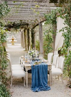 an outdoor dining table set with blue linens and white chairs, surrounded by greenery