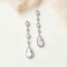 a pair of white diamond drop earrings on top of a furnishing cloth with feathers in the background