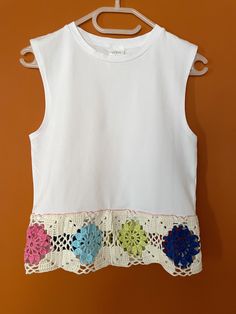 a white top with crocheted flowers on the front and bottom, hanging from a wooden hanger