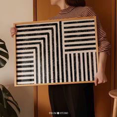 a woman holding up a black and white framed art piece in front of a plant