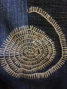 an embroidered spiral on the back pocket of a pair of blue denim jeans with gold thread