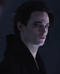 a young man with dark hair and blue eyes looks into the distance while wearing a black coat