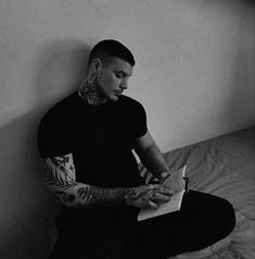 a man with tattoos sitting on a bed holding a book and looking at the camera