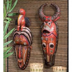 two african masks sitting next to each other on a wooden surface with plants in the background